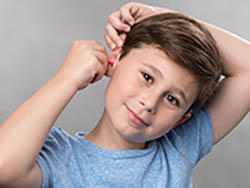 A boy uses two hands to pull the top of his ear up and back and slides a formable foam earplug into his ear.