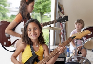 Young children in a garage playing guitars and drums.