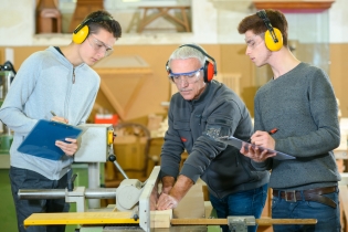 Estudiantes de secundaria y un maestro, que está usando una herramienta eléctrica, usando orejeras en una clase de carpintería.