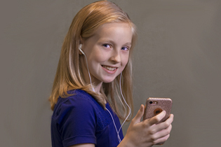 A preteen girl holds a smartphone in her hand and listens to music through earbuds.