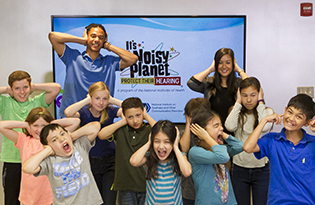Two educators and about a dozen students, approximately ages 8 to 12, hold their hands over the ears as if the room is very loud. A slide from the Noisy Planet Teacher Toolkit is visible on a large screen behind the group.