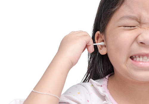 A young girl wincing as she cleans her ear with a cotton swab.