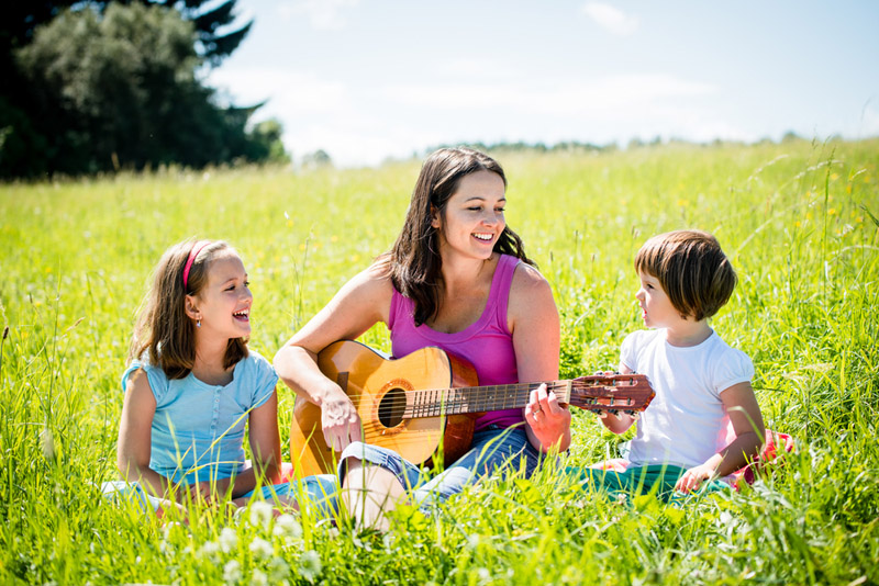 A mother teaches her children to play guitar in a field on a sunny day