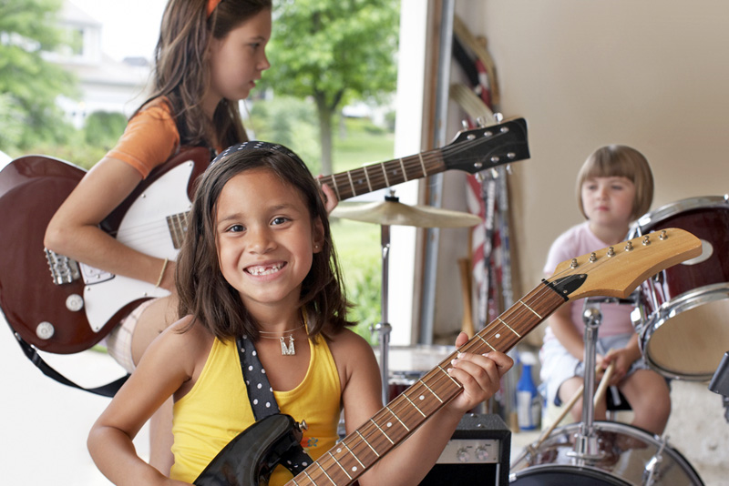 Young children in a garage playing guitars and drums.
