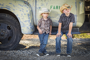 Two young boys wearing cowboy hats leaning against an antique truck in a rustic country setting.