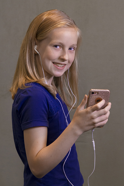 A preteen girl holds a smartphone in her hand and listens to music through earbuds.