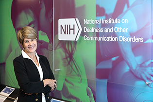 An image of Dr. Tucci standing in a hallway next to a sign that reads, 'NIH National Institute on Deafness and Other Communication Disorders.'
