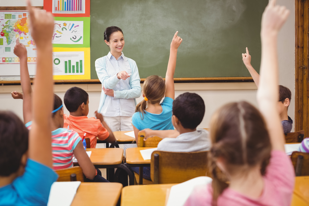 Teacher asking her elementary students a question in a classroom. Students are raising their hands to answer the question.