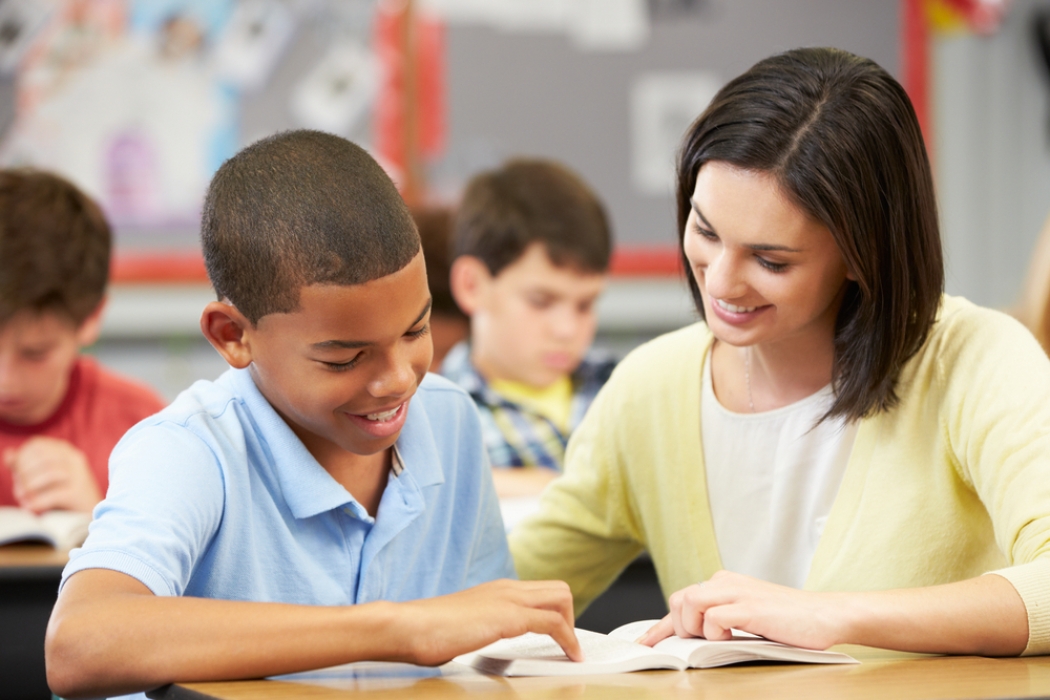 Una maestra sentada con un estudiante en un salón escolar leyendo un libro.