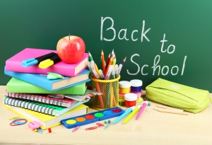 Books, pencils, pens, paperclips, craft paint, pencil case, and an apple are on a desk in front of a chalkboard with “Back to School” written on it.