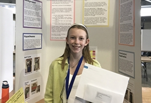 13-year-old Nora Keegan stands in front of a  poster presentation detailing her research.