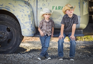 Two young boys wearing cowboy hats lean against an antique truck in a rustic country setting.