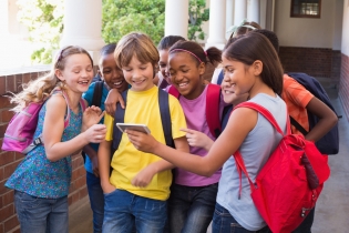 A group of children standing in the hallway of a school looking at a mobile device.