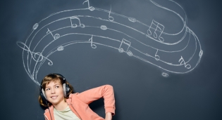 Una niña usando audiófonos mientras escucha su reproductor de música. Está delante de una pizarra con notas musicales escritas con gis o tiza.