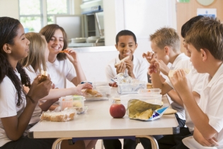 Niños en un colegio sentados alrededor de la mesa hablando y comiendo su almuerzo.