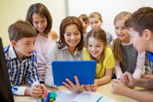 Una maestra y un grupo de niños están sentados en un salón de clase mirando una computadora tipo tableta. 