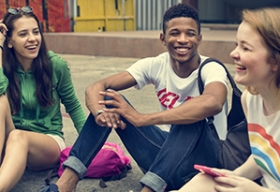 Three teenagers sitting on the ground laughing and talking with each other.