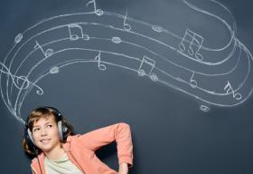 Una niña usando audiófonos mientras escucha su reproductor de música. Está delante de una pizarra con notas musicales escritas con gis o tiza.