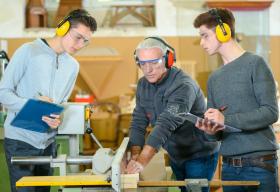 High school students and a teacher, who is using a power tool, wearing protective earphones in a woodworking class.