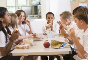Niños en un colegio sentados alrededor de la mesa hablando y comiendo su almuerzo.